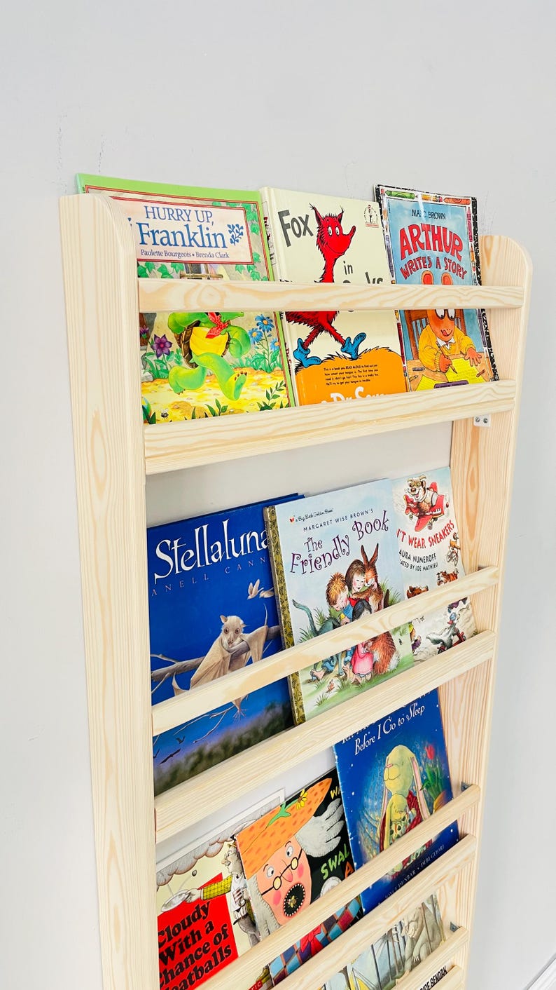 May include: A wooden bookshelf filled with colorful children's books. Titles include "Hurry Up, Franklin", "Fox in Socks", "Arthur Writes a Story", and "Stellaluna". The bookshelf is made of light-colored wood and is mounted on a white wall.