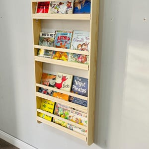 May include: A wall-mounted, light-colored wooden bookshelf filled with children's books. The books have colorful covers and titles such as "Dogs Don't Wear Sneakers", "Arthur Writes a Story", and "The Night Pirates". The bookshelf is mounted on a light gray wall.