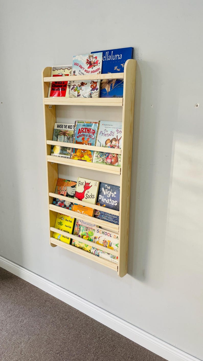 May include: A wall-mounted, light-colored wooden bookshelf filled with children's books. The books have colorful covers and titles such as "Dogs Don't Wear Sneakers", "Arthur Writes a Story", and "The Night Pirates". The bookshelf is mounted on a light gray wall.