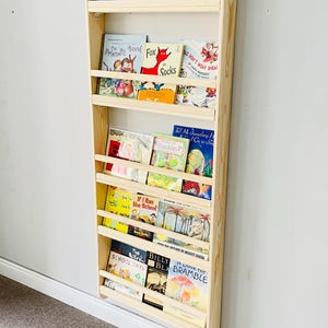 May include: A wooden bookshelf with multiple shelves, filled with colorful children's books. The bookshelf is mounted on a white wall. Titles include "Stellaluna", "Arthur Writes a Story", and "The Friendly Book".