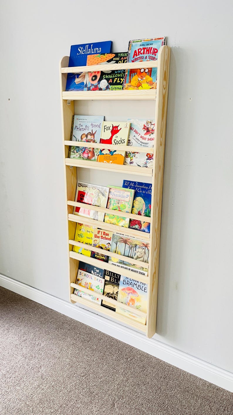 May include: A wooden bookshelf with multiple shelves, filled with colorful children's books. The bookshelf is mounted on a white wall. Titles include "Stellaluna", "Arthur Writes a Story", and "The Friendly Book".
