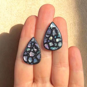 Könnte beinhalten: Zwei tropfenförmige Ohrringe mit schwarzer Basis und eingelegten irisierenden Teilen. Die Ohrringe werden auf einer Hand präsentiert und zeigen das farbenfrohe, mosaikartige Design. Die Ohrringe haben eine polierte Oberfläche und fangen das Licht ein.