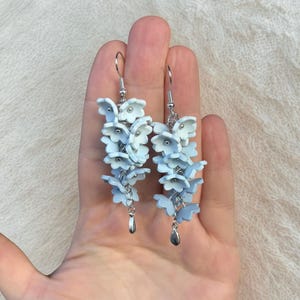 Könnte beinhalten: Ein Paar hellblaue Blumenohrringe mit silberfarbenen Haken und tropfenförmigen Anhängern. Die Ohrringe zeigen Cluster aus kleinen, zarten Blüten in verschiedenen Blau- und Weißtönen, die einen Kaskadeneffekt erzeugen. Die Ohrringe sind zum Hängen gedacht.