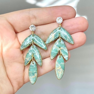 May include: A pair of dangle earrings featuring three tiered, leaf-shaped segments in a marbled seafoam green finish with gold-toned veining. Each earring is topped with a single clear, round faceted crystal stud set in a gold-toned metal frame.