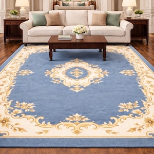 Puede incluir: Una alfombra azul con un borde floral crema y dorado y un medallón central. La alfombra está colocada en un salón con un sofá blanco, una mesa de centro de madera y mesas auxiliares a juego.