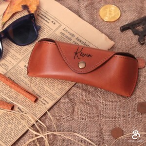 May include: A brown leather sunglasses case with the name "Kewin" in script. The case is closed with a snap and sits on a vintage newspaper. A pair of sunglasses and a gold coin are also visible.