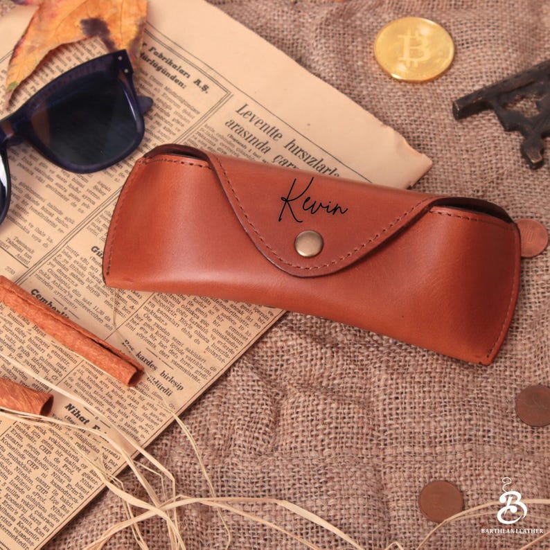 May include: A brown leather sunglasses case with the name "Kewin" in script. The case is closed with a snap and sits on a vintage newspaper. A pair of sunglasses and a gold coin are also visible.