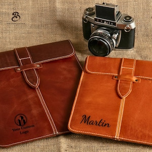 May include: Two leather tablet cases in shades of brown and tan with white stitching and a flap closure. One case is embossed with "Your Custom Logo", the other with the name "Martin". A vintage camera sits in the background.
