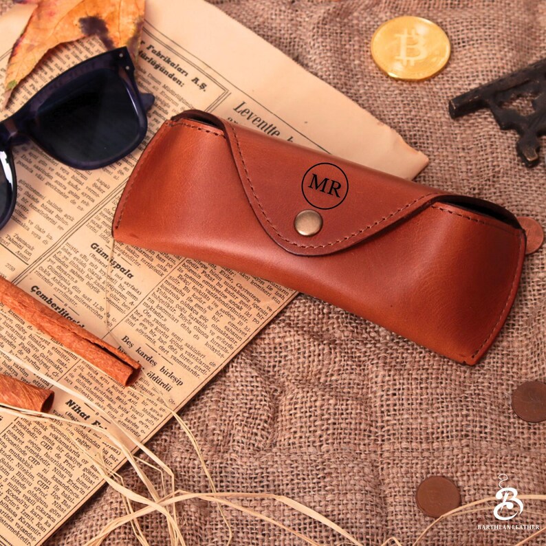 Puede incluir: Un estuche de gafas de cuero marr&oacute;n con las iniciales "MR" grabadas en la solapa. El estuche se cierra con un broche y se encuentra sobre una superficie de arpillera con un peri&oacute;dico y gafas de sol.