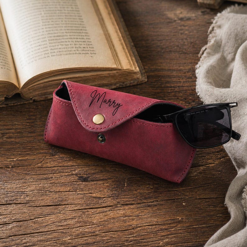 Puede incluir: Un estuche de gafas de cuero burdeos con la palabra "Marry" en cursiva. El estuche est&aacute; abierto, revelando un par de gafas de sol negras. El estuche tiene un cierre de bot&oacute;n a presi&oacute;n y un bot&oacute;n dorado. El fondo incluye un libro abierto y un pa&ntilde;o de lino.