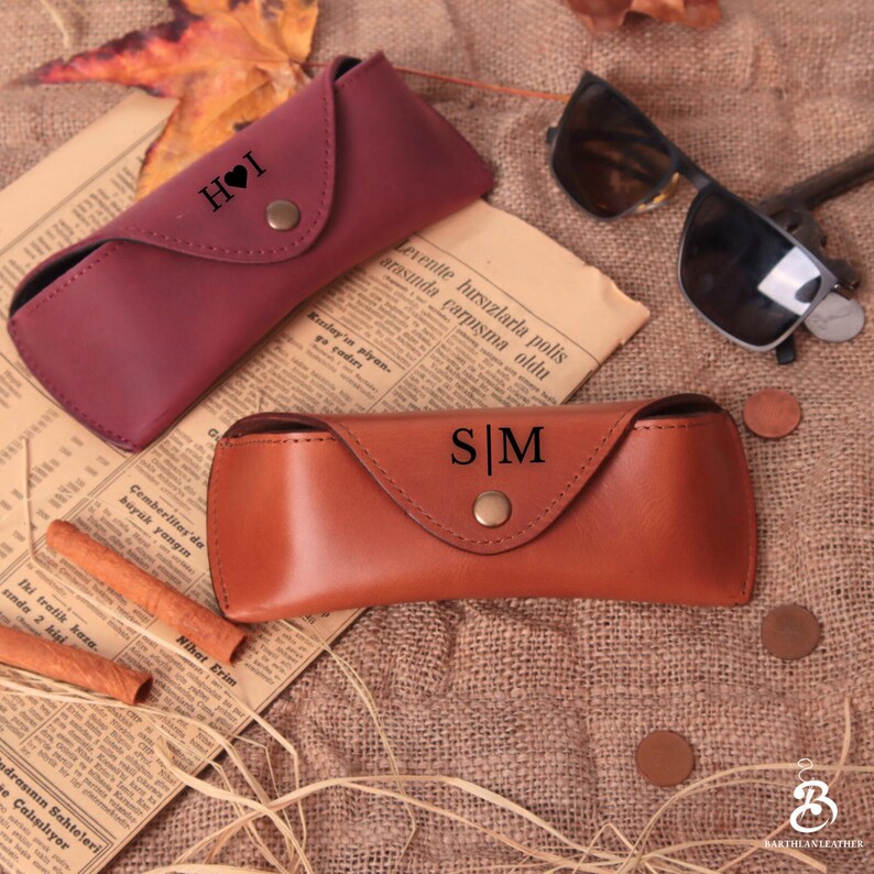 Puede incluir: Dos estuches de cuero para gafas, uno burdeos y otro marr&oacute;n, con iniciales personalizadas. El estuche burdeos tiene "H I" con un s&iacute;mbolo de coraz&oacute;n, y el estuche marr&oacute;n tiene "S | M". Gafas de sol y recortes de peri&oacute;dico en el fondo.