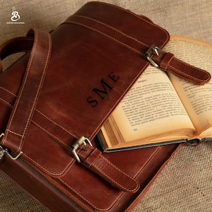 May include: A brown leather messenger bag with the initials "SME" embossed on the front flap. The bag features a shoulder strap, buckle closures, and visible stitching. An open book is inside the bag. The bag is made of leather.