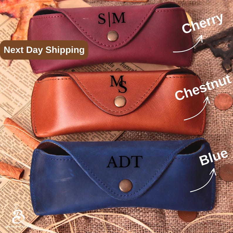 May include: Three leather eyeglass cases in cherry, chestnut, and blue. Each case has a snap closure and personalized initials. The cherry case has "S|M", the chestnut case has "M", and the blue case has "ADT".
