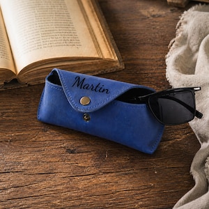 Puede incluir: Un estuche de gafas de cuero azul con el nombre "Martin" grabado en relieve en la parte delantera. El estuche est&aacute; abierto, revelando un par de gafas de sol negras. El estuche tiene un cierre de bot&oacute;n a presi&oacute;n y un bot&oacute;n dorado. El fondo incluye un libro abierto y una superficie de madera.