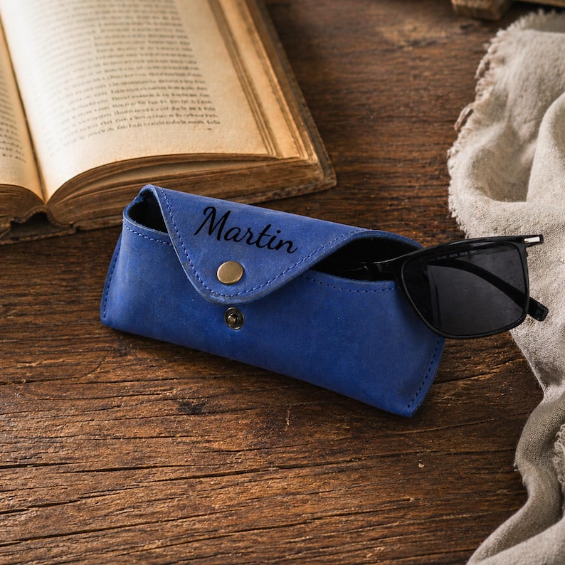 Puede incluir: Un estuche de gafas de cuero azul con el nombre "Martin" grabado en relieve en la parte delantera. El estuche est&aacute; abierto, revelando un par de gafas de sol negras. El estuche tiene un cierre de bot&oacute;n a presi&oacute;n y un bot&oacute;n dorado. El fondo incluye un libro abierto y una superficie de madera.
