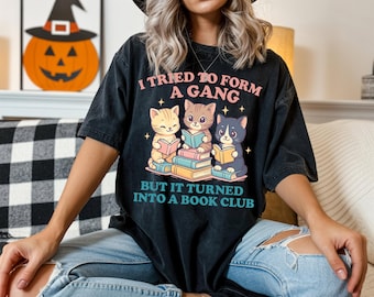 May include: Black t-shirt with the text "I tried to form a gang but it turned into a book club." The shirt features a graphic of three cartoon cats reading books. The cats are sitting on a stack of books.