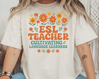 May include: A cream-coloured t-shirt featuring the text "ESL TEACHER" in orange, and "CULTIVATING LANGUAGE LEARNERS" in teal. The design includes floral elements in orange, yellow, and green. A casual, comfortable style, ideal for teachers.