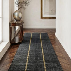 May include: A long, rectangular runner rug featuring a dark charcoal gray woven texture with two prominent, parallel vertical stripes in a muted gold tone. The rug is finished with short, frayed fringe detailing along both of its narrow ends.