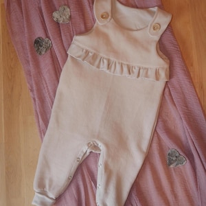 Könnte beinhalten: Ein hellbeige Baby-Strampler mit Waffelstruktur, mit Rüschendetail an der Brust und Knopfverschlüssen. Der Strampler hat einen runden Ausschnitt und ist mit zwei Holzknöpfen an den Schultern versehen. Das Kleidungsstück wird auf einem rosa Stoff präsentiert.
