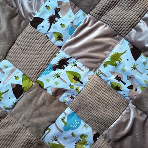 Könnte beinhalten: Eine Patchwork-Babydecke mit Quadraten aus verschiedenen Texturen und Mustern. Das Design umfasst hellblauen Stoff mit Cartoon-Dinosauriern, grünen Blättern und Palmen. Weitere Quadrate sind aus braunem Waffelgewebe, Plüsch und Satin.
