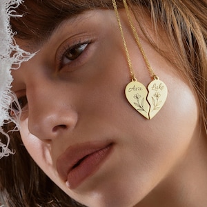 Puede incluir: Un collar dorado con un colgante de corazón roto. Cada mitad del corazón está grabada con un nombre y un diseño floral. Los nombres "Ava" y "Luke" son visibles en el colgante.