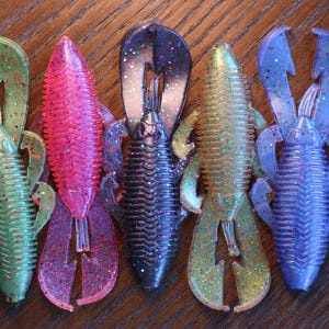 May include: Five colorful plastic fishing lures in shades of green, pink, black, gold, and blue. Each lure has a segmented body and a claw-like tail, with glitter accents. The lures are arranged on a wooden surface.