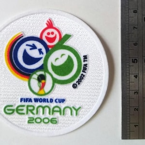 Könnte beinhalten: Ein kreisförmiger, weißer Stickaufnäher mit dem Logo der FIFA Fußball-Weltmeisterschaft Deutschland 2006, bunten lachenden Symbolen und Text. Ein Metalllineal zeigt, dass der Aufnäher einen Durchmesser von etwa 8,3 cm hat.