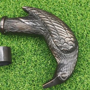 May include: A dark bronze-colored cane handle shaped like a bird's head, with detailed feather carvings. Includes a metal ring and a silver screw, all resting on a green surface. The handle is likely for a walking stick or similar item.
