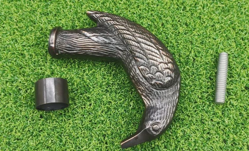 May include: A dark bronze-colored cane handle shaped like a bird's head, with detailed feather carvings. Includes a metal ring and a silver screw, all resting on a green surface. The handle is likely for a walking stick or similar item.