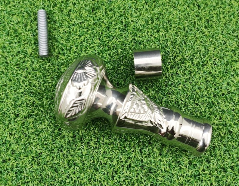 May include: A silver-colored cane handle with floral and bird-like designs. The handle is accompanied by a silver metal cylinder and a silver screw. The items are displayed on a green surface.