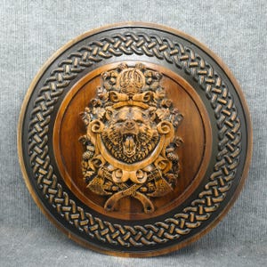 May include: Circular wooden wall art with a carved bear's head, open-mouthed, crowned, and surrounded by axes and foliage. The bear is central, framed by a Celtic knot design. The piece is made of wood.
