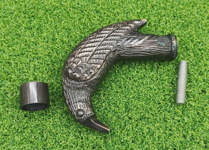 May include: A bronze-colored cane handle shaped like a bird's head, with detailed feather carvings. Includes a metal collar and a silver screw. The handle is approximately 6 inches long.