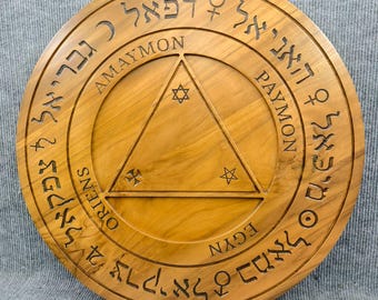 May include: A circular wooden plaque with engraved text and symbols. The plaque features a central triangle with stars and crosses, surrounded by circular text. The wood has a warm, natural tone, and the engraved details are dark.