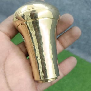 May include: A polished brass knob with a smooth, rounded top featuring subtle horizontal grooves and a slightly tapered, hammered-texture body. The metallic surface reflects light, showcasing a warm, golden tone and a cylindrical base.