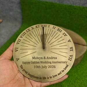 Op de afbeelding: Een ronde zonnewijzer van messing met Romeinse cijfers van I tot XII langs de rand en een centrale verticale gnomon. De gegraveerde tekst luidt: Melvyn & Andrea, Happy Golden Wedding Anniversary, 10th July 2026, The great happiness in life is to love and be loved.