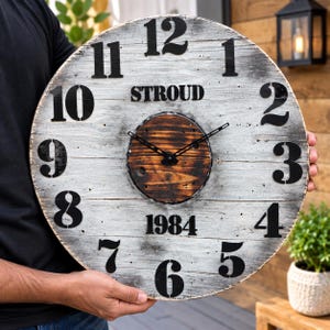 May include: A large round wall clock made of weathered white wooden planks with black painted numbers from 1 to 12. The center features a dark stained circular wood panel with black metal hands. The text STROUD and 1984 are printed in black.