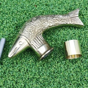 Puede incluir: Un mango de bastón de latón con diseño de pájaro, un tornillo de metal y un anillo de latón se muestran sobre una superficie verde. El mango del pájaro presenta intrincados detalles grabados. Los componentes parecen estar separados.