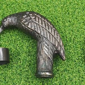 May include: A detailed, bronze-colored cane handle shaped like a bird's head, with intricate feather and wing detailing. Includes a matching ferrule and a silver-colored screw. Set against a green, grassy background.