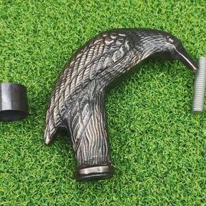 May include: A detailed, dark metal eagle head cane handle with a textured feather design. Includes a separate metal ring and a threaded metal rod. The handle is set against a green, grassy background.