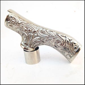 May include: A silver-coloured, ornate cane handle with a floral pattern. The handle is curved and has a cylindrical base. The design is detailed and appears to be made of metal.