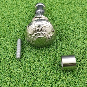 May include: A silver-colored, decorative furniture knob with a detailed floral design. The knob is accompanied by a threaded bolt and a small metal sleeve, all resting on a green surface.