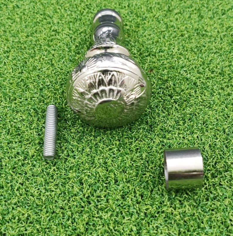 May include: A silver-colored, decorative furniture knob with a detailed floral design. The knob is accompanied by a threaded bolt and a small metal sleeve, all resting on a green surface.