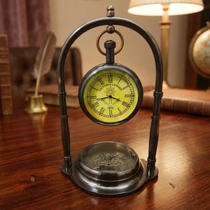 May include: A dark metal desk clock suspended within an arched frame, featuring a yellow face with Roman numerals, an anchor graphic, and the text "VICTORIA LONDON 1876". A circular compass is integrated into the base of the stand.