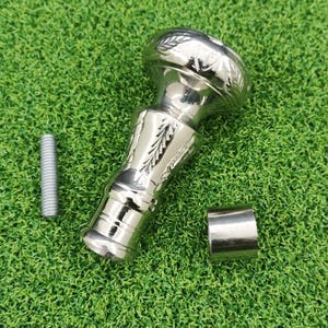 May include: A silver-colored cane handle with a decorative floral design. The handle is accompanied by a silver metal ring and a silver screw. The items are on a green surface.