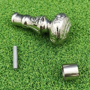May include: A silver-colored walking cane handle with an ornate design featuring an eagle and floral patterns. The handle is accompanied by a silver screw and a silver metal ring. The items are on a green surface.