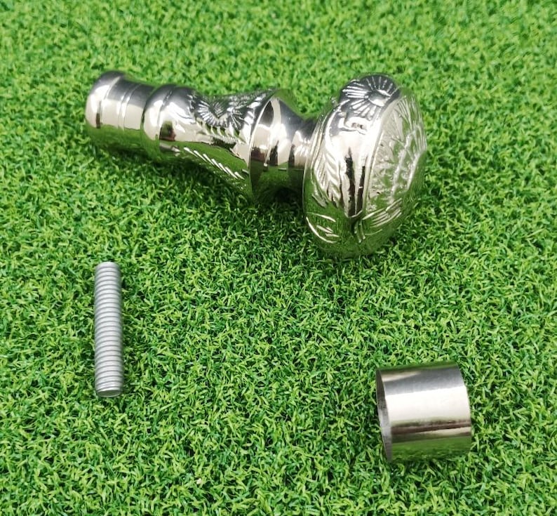 May include: A silver-colored walking cane handle with an ornate design featuring an eagle and floral patterns. The handle is accompanied by a silver screw and a silver metal ring. The items are on a green surface.