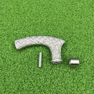 May include: A silver-coloured cane handle with a woven pattern, a screw, and a metal ferrule. The handle is curved and has a textured surface. The items are on a green surface.
