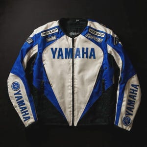May include: A Yamaha motorcycle racing jacket in blue, white, and black. The jacket features the Yamaha logo across the chest and on the sleeves, with Dunlop and Ohlins patches. The jacket has a zippered front and a stand-up collar.