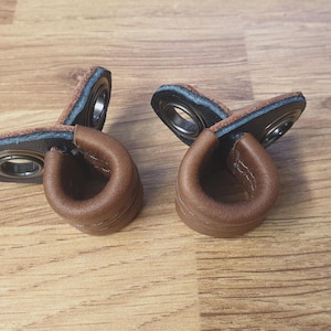 May include: Two brown leather drawer pulls with a metal bearing and a stitched design. The pulls are in a U-shape, with a dark blue and brown backing. The background is a light brown wooden surface.