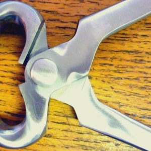 Könnte beinhalten: Nahaufnahme eines silbernen Hufschneiders. Das Werkzeug hat gebogene Backen und lange Griffe, die zum Beschneiden von Pferdehufeisen bestimmt sind. Das Metallwerkzeug ist vor einem Holzgrund platziert.